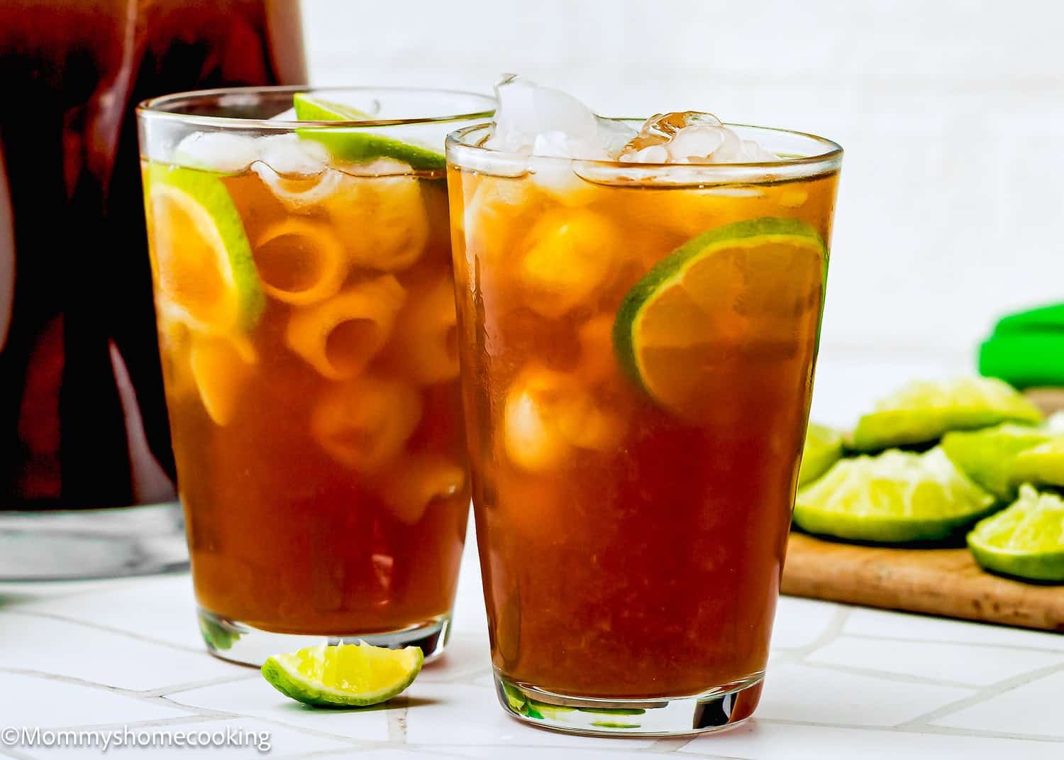 Two glasses of iced papelon con Limon with ice cubes and lime slices on a white surface, with lime wedges and a pitcher in the background.