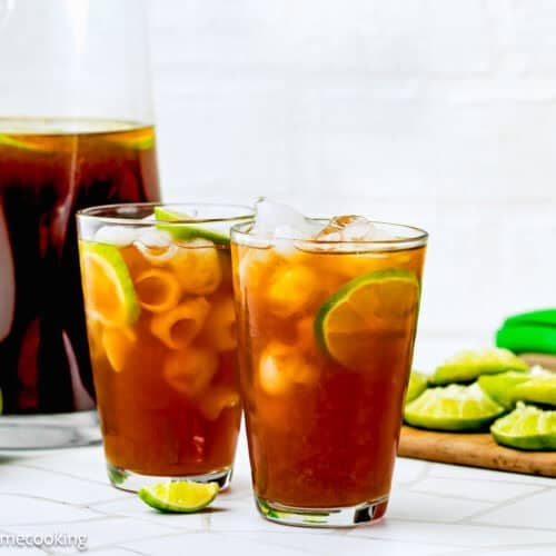 Two glasses of iced papelon con limon with lime slices and ice cubes, a pitcher of papelon con limon, whole and sliced limes, a green citrus squeezer, on a white surface.