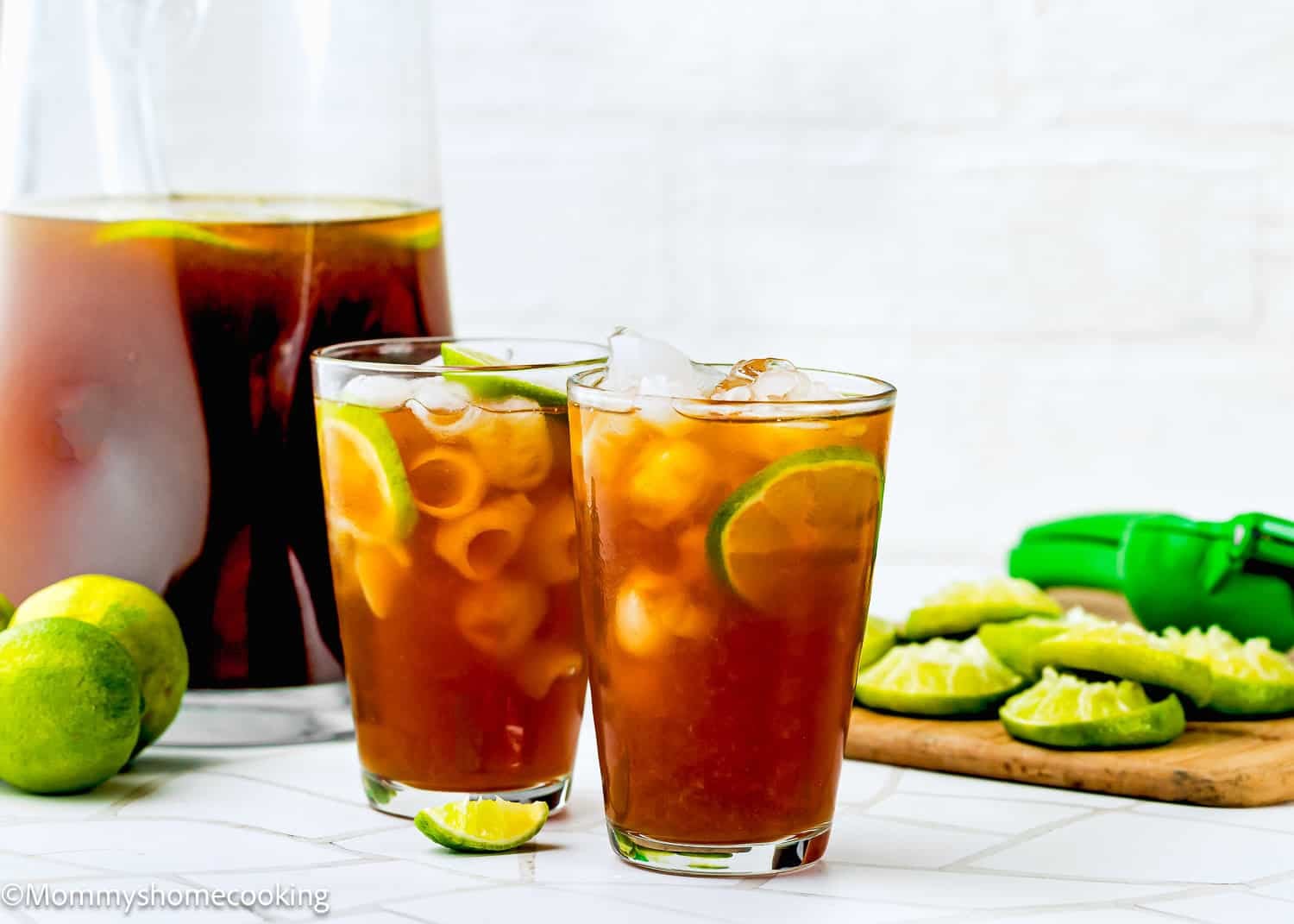 Two glasses of iced papelon con Limon with lime slices and ice cubes, a pitcher of papelon con Limon, whole and sliced limes, a green citrus squeezer, on a white surface.