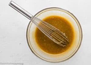A metal whisk rests in a glass bowl filled with a light brown mixture for Egg-Free Sheet Pan Chocolate Chip Cookies on a white surface.