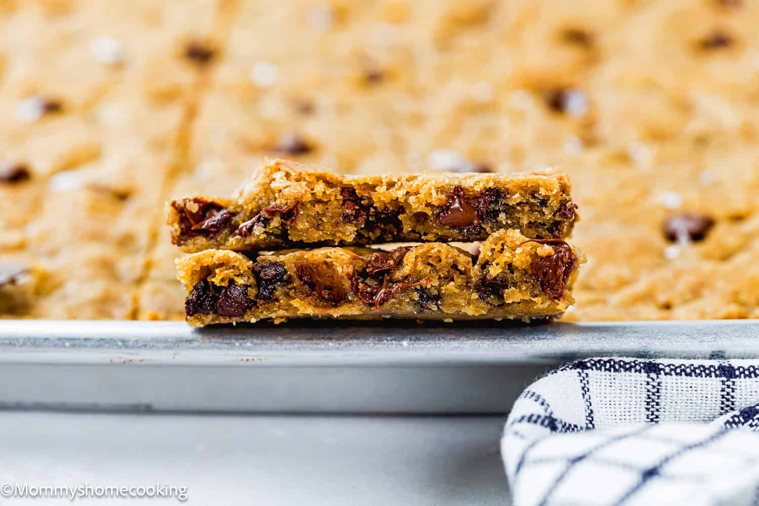 Two stacked Egg-Free Sheet Pan Chocolate Chip Cookies rest on the edge of a baking sheet with more cookie bars in the background, next to a striped towel.