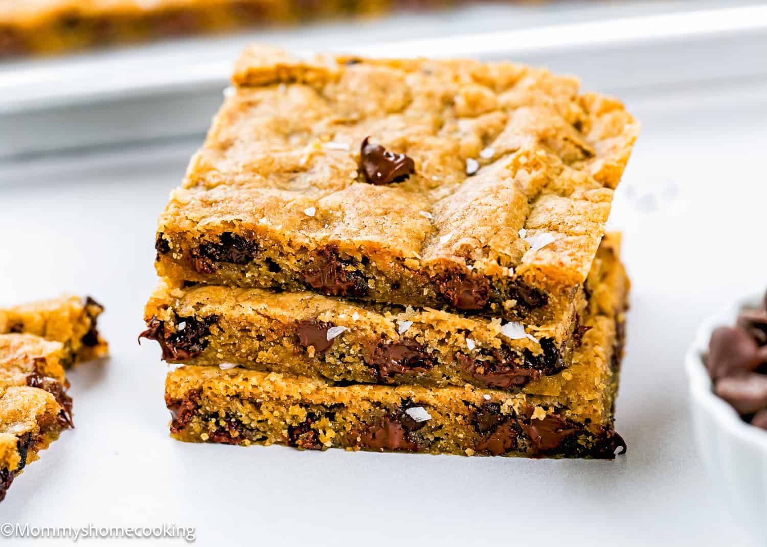 Three stacked Egg-Free Sheet Pan Chocolate Chip Cookie bars on a white surface, with visible chocolate chips and a sprinkle of sea salt on top.