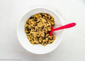 A white bowl filled with egg-free sheet pan chocolate chip cookie dough and a red spatula resting inside, placed on a light-colored surface.