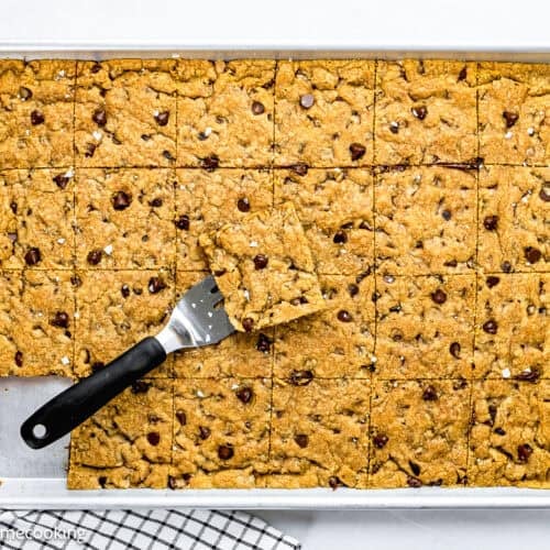 A baking sheet with Egg-Free Sheet Pan Chocolate Chip Cookie bars cut into squares, one piece lifted with a spatula. A checkered kitchen towel and some chocolate chips are nearby.