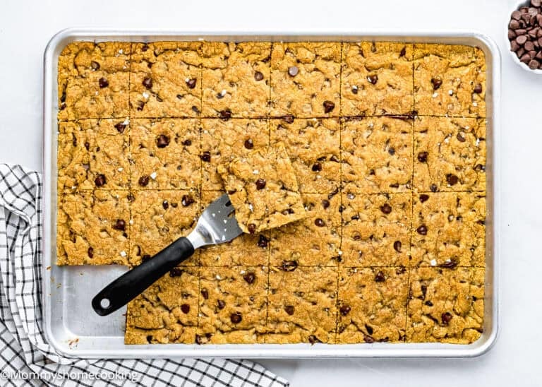A baking sheet with Egg-Free Sheet Pan Chocolate Chip Cookie bars cut into squares, one piece lifted with a spatula. A checkered kitchen towel and some chocolate chips are nearby.