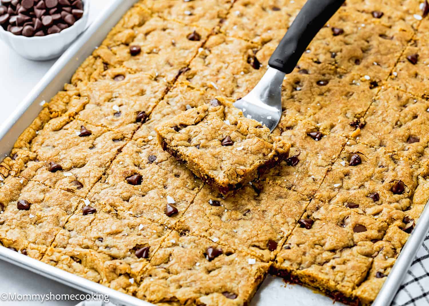 A baking sheet of Egg-Free Sheet Pan Chocolate Chip Cookies cut into squares, with one square being lifted by a spatula.