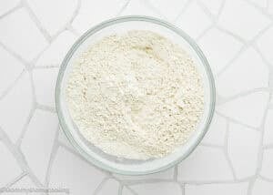 A glass bowl filled with flour and a dry baking mixture for Eggless Banana Bars With Cream Cheese Frosting sits on a white tiled surface.