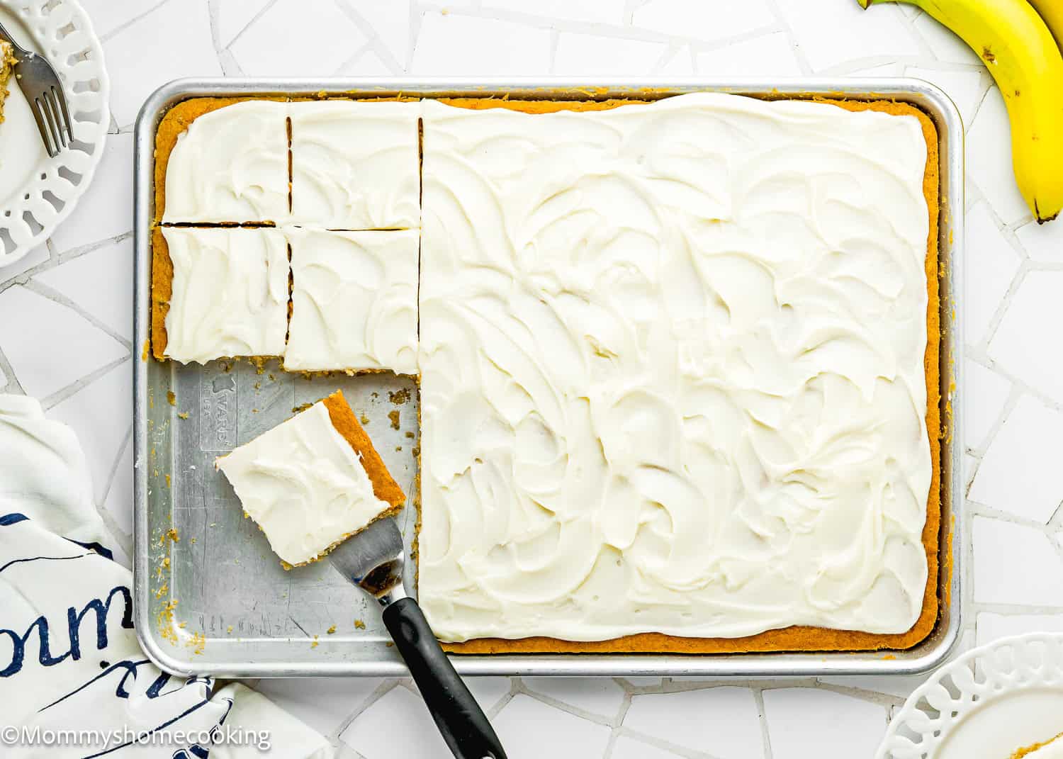 A sheet pan of Eggless Banana Bars With Cream Cheese Frosting, with six square pieces cut and one piece being lifted out with a spatula; bananas and plates are nearby.