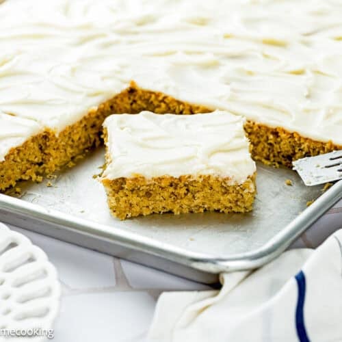 A frosted sheet cake of Eggless Banana Bars With Cream Cheese Frosting has one square piece cut out and placed in front on a metal baking sheet, with a serving spatula and a decorative plate nearby.
