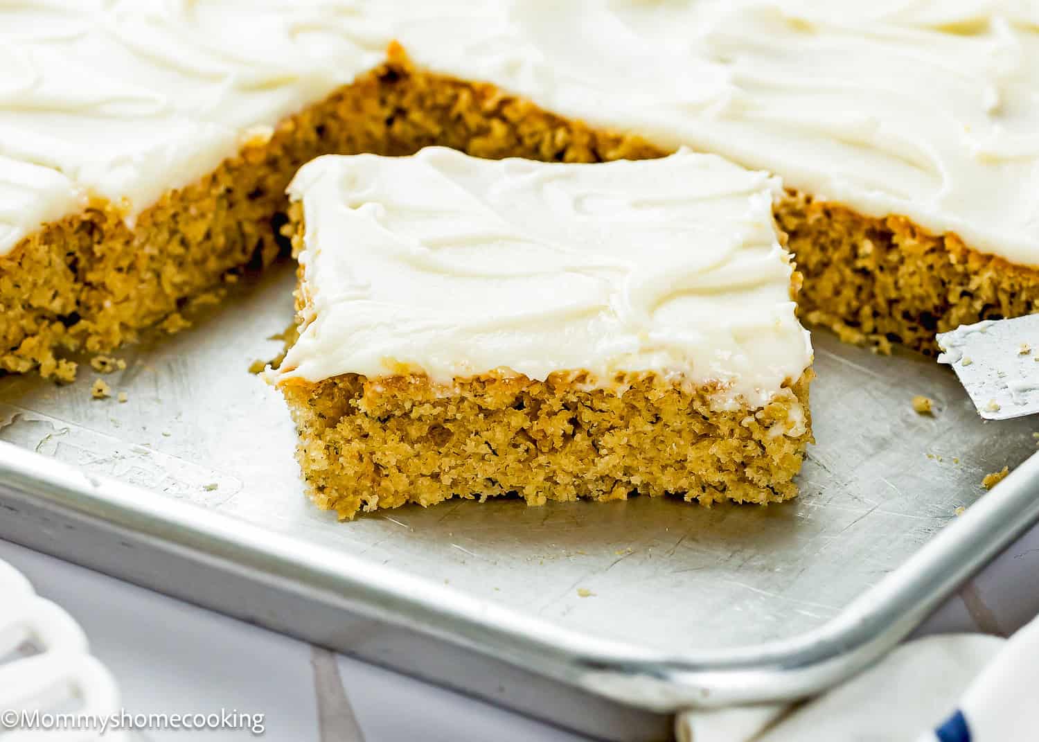 A sliced sheet cake of Eggless Banana Bars With Cream Cheese Frosting sits on a metal baking tray, with one piece slightly separated from the rest.