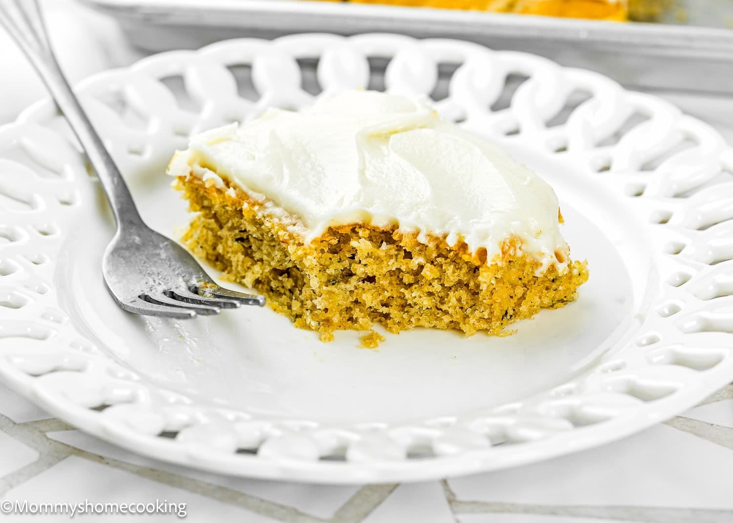 A slice of Eggless Banana Bars With Cream Cheese Frosting, with a bite taken out, served on a white decorative plate with a fork.