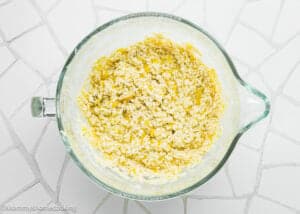 Glass mixing bowl with a yellow and white crumbly dough mixture for Eggless Banana Bars With Cream Cheese Frosting, placed on a white patterned surface.