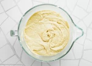 A glass mixing bowl filled with creamy, light yellow batter for Eggless Banana Bars With Cream Cheese Frosting sits on a white patterned surface.
