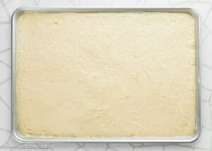 A metal baking sheet filled with an even layer of uncooked Eggless Banana Bars With Cream Cheese Frosting batter sits on a white patterned surface.