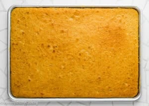 A rectangular sheet pan filled with evenly baked, golden Eggless Banana Bars With Cream Cheese Frosting on a white, patterned surface.