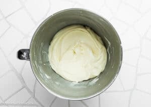 A mixing bowl filled with smooth, creamy white frosting for Eggless Banana Bars With Cream Cheese Frosting sits on a white tiled surface.