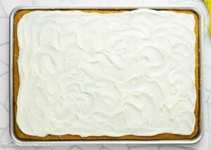 A rectangular sheet cake, Eggless Banana Bars With Cream Cheese Frosting, features white frosting covering the entire surface and is displayed on a metal baking tray.