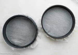 Two round cake pans lined with parchment paper, ready for making an Eggless Chocolate Chip Cookie Ice Cream Cake, placed side by side on a light-colored surface.