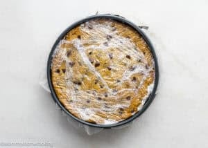 A round baking pan filled with Eggless Chocolate Chip Cookie Ice Cream Cake dough, covered with plastic wrap, sits on a white surface.
