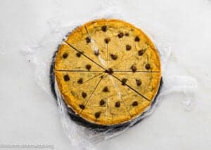 An Eggless Chocolate Chip Cookie Ice Cream Cake, round and cut into twelve slices, sits on plastic wrap atop a white surface.