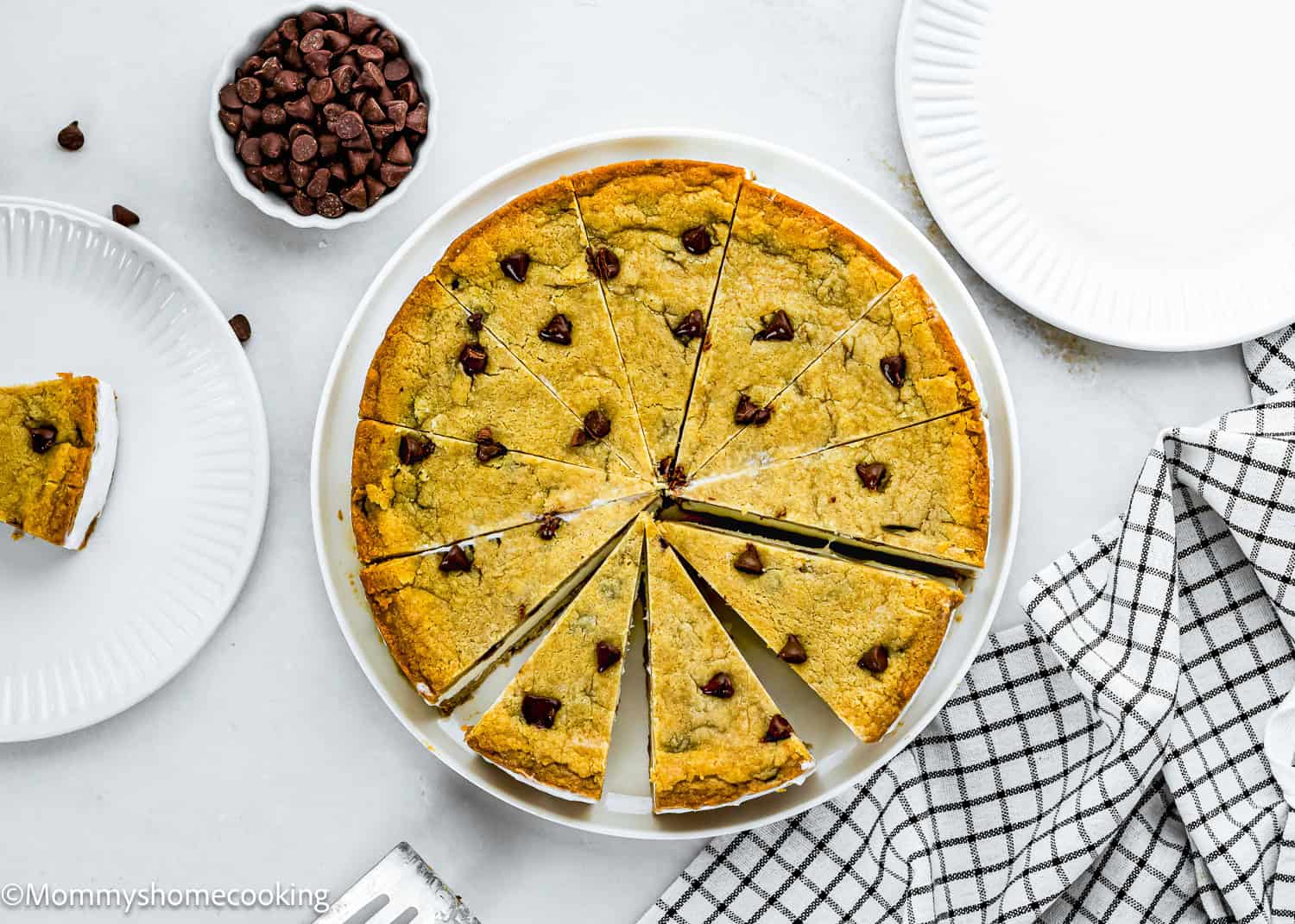 A large Eggless Chocolate Chip Cookie Ice Cream Cake cut into slices is displayed on a white plate, with a slice served on a side plate, a bowl of chocolate chips, and a checkered towel nearby.