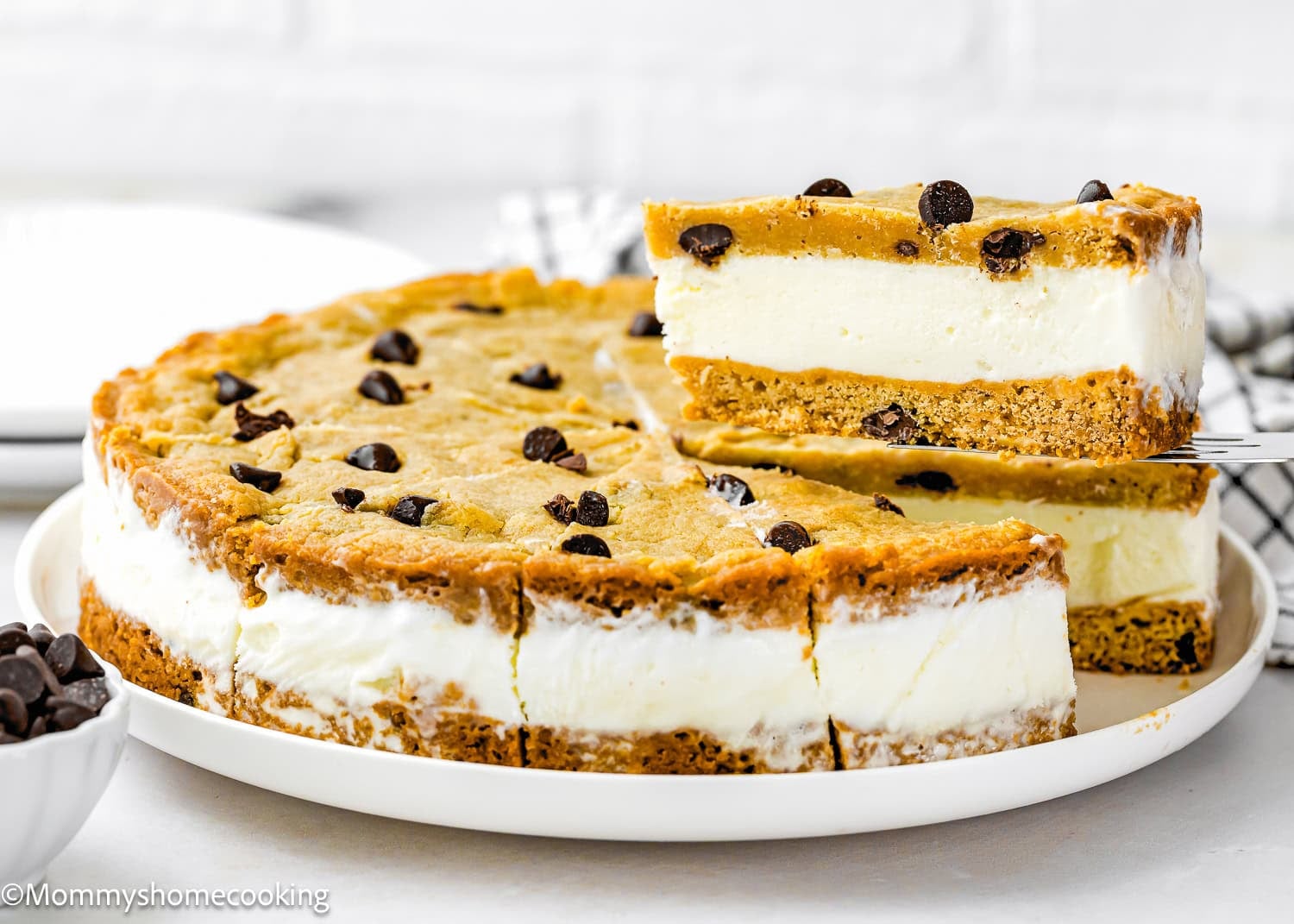 A slice of Eggless Chocolate Chip Cookie Ice Cream Cake is being lifted from a round cake, revealing delicious layers of cookie and vanilla ice cream.