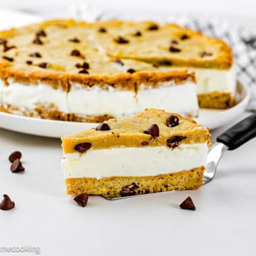 A slice of Eggless Chocolate Chip Cookie Ice Cream Cake is being served from a larger cake, with chocolate chips scattered nearby.