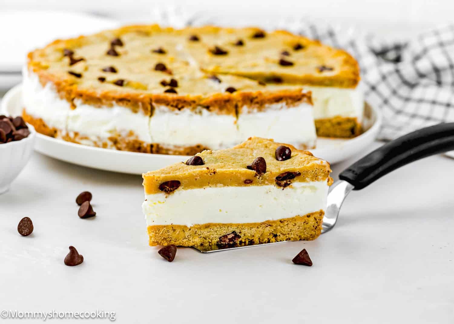 A slice of Eggless Chocolate Chip Cookie Ice Cream Cake is being served from a larger cake, with chocolate chips scattered nearby.