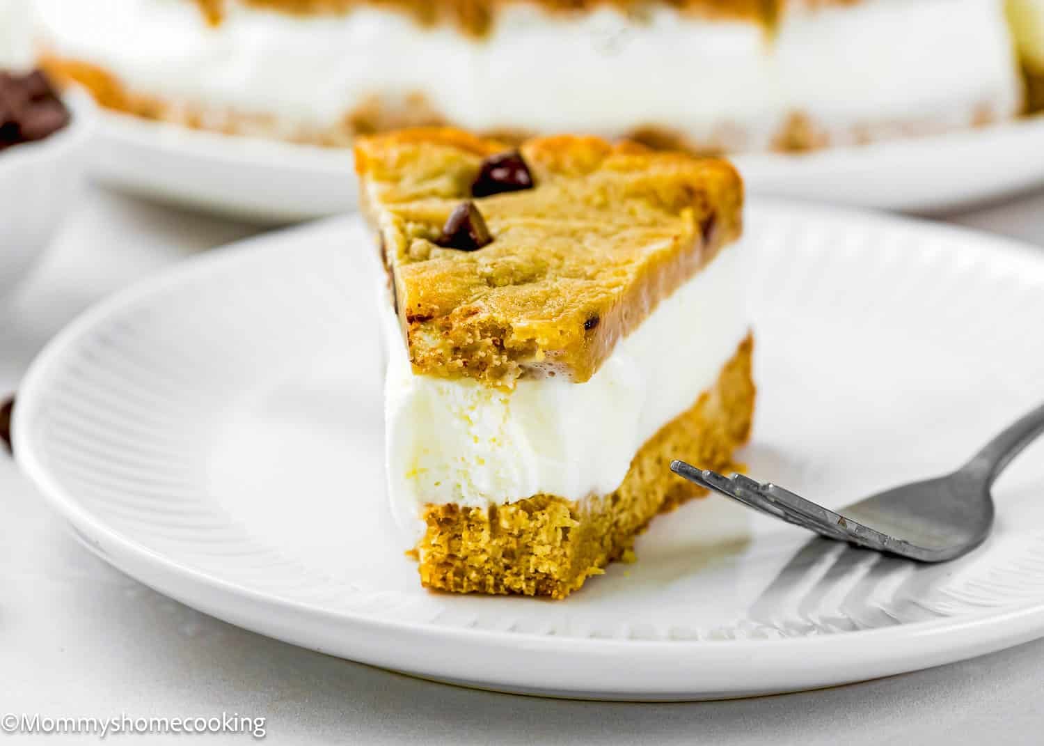 A slice of Eggless Chocolate Chip Cookie Ice Cream Cake with a cookie crust and chocolate chips on a white plate, with a fork beside it.