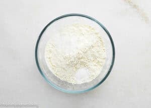A clear glass bowl filled with a mixture of dry white powders, possibly for making Eggless Chocolate Chip Cookie Ice Cream Cake, sits on a light-colored surface.