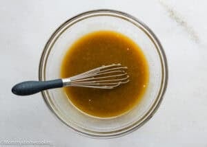 A glass bowl with a metal whisk mixes a brown liquid sauce or dressing, perfect for Eggless Chocolate Chip Cookie Ice Cream Cake, on a white surface.