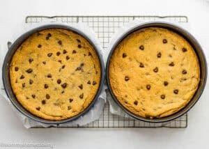 Two round pans of Eggless Chocolate Chip Cookie Ice Cream Cake cooling on a wire rack; the left has more chocolate chips visible on top than the right.