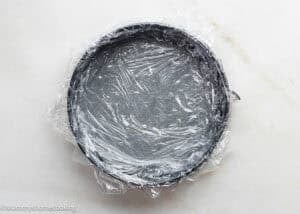A round baking pan lined with plastic wrap, viewed from above on a white surface, ready for making an Eggless Chocolate Chip Cookie Ice Cream Cake.