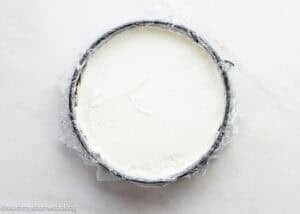 A round pan lined with plastic wrap holds a smooth layer of white cream cheese mixture, perfect as the base for an Eggless Chocolate Chip Cookie Ice Cream Cake, viewed from above.