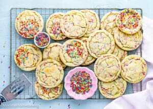 A cooling rack holds several large Eggless Drop Sugar Cookies with Sprinkles, surrounded by bowls of extra sprinkles on a light blue surface.