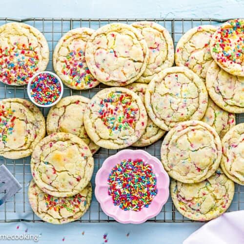 A cooling rack holds several large Eggless Drop Sugar Cookies with Sprinkles, surrounded by bowls of extra sprinkles on a light blue surface.