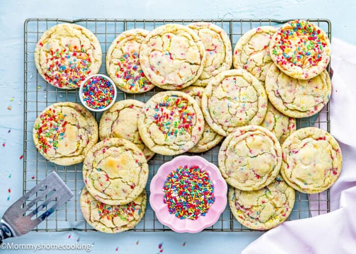 A cooling rack holds several large Eggless Drop Sugar Cookies with Sprinkles, surrounded by bowls of extra sprinkles on a light blue surface.
