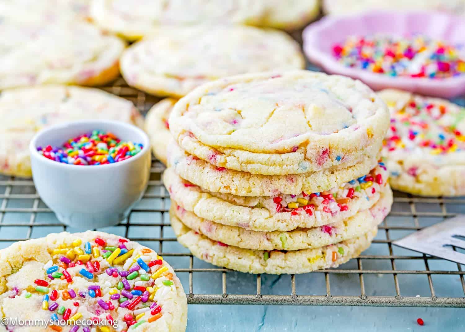 A stack of Eggless Drop Sugar Cookies with Sprinkles sits on a cooling rack, surrounded by more cookies and a small bowl of sprinkles.