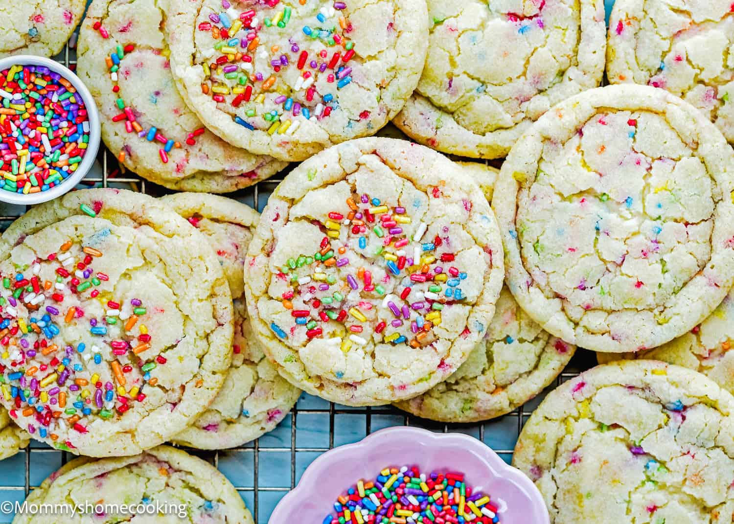 A batch of Eggless Drop Sugar Cookies with Sprinkles cooled on a rack, surrounded by small bowls of extra sprinkles.