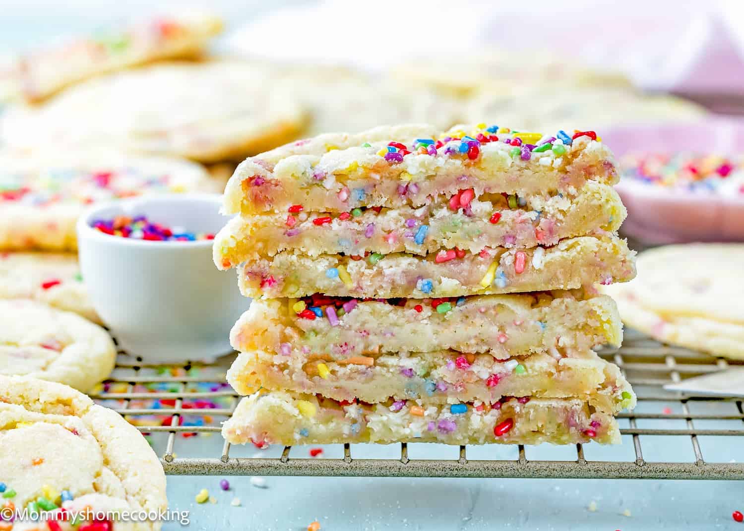 Stack of Eggless Drop Sugar Cookies with Sprinkles, broken in half to show a soft interior, on a cooling rack with more cookies and a small bowl of sprinkles in the background.