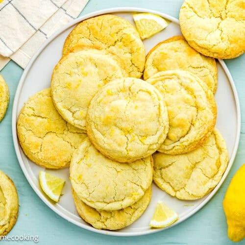 A plate of Eggless Lemon Sugar Cookies (Soft & Chewy) with lemon wedges, a whole lemon, and a striped towel on a light blue surface.