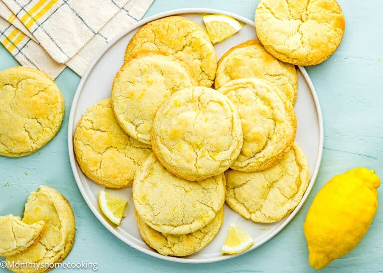 A plate of Eggless Lemon Sugar Cookies (Soft & Chewy) with lemon wedges, a whole lemon, and a striped towel on a light blue surface.