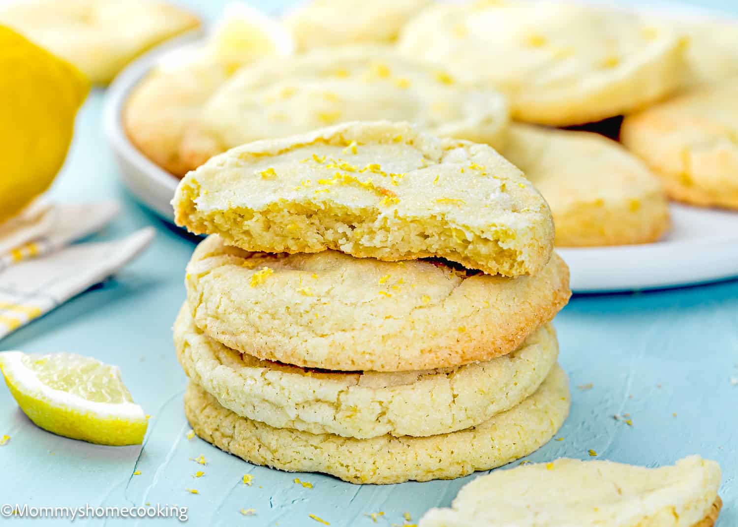 A stack of Eggless Lemon Sugar Cookies (Soft & Chewy), with one broken in half on top, sits on a light blue surface. In the background, a plate holds more cookies and fresh lemon slices.