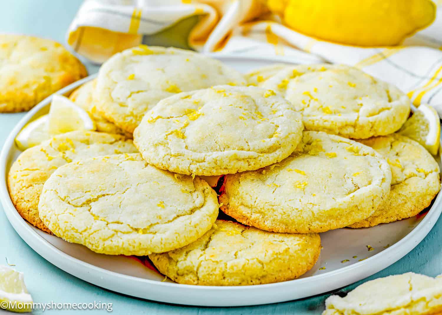 A plate of Eggless Lemon Sugar Cookies (Soft & Chewy) piled on top of each other, with a yellow and white cloth and lemon slices in the background.
