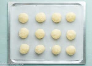 A baking tray lined with parchment paper holds twelve evenly spaced, round, unbaked dough balls coated in sugar for Eggless Lemon Sugar Cookies (Soft & Chewy).