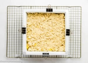 Square baking pan lined with parchment paper and filled with crumb topping for Eggless Peach Crumb Bars, secured with binder clips, sits on a cooling rack.