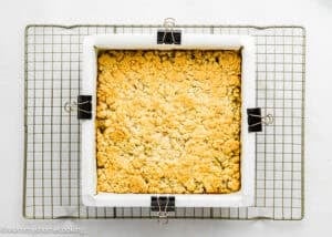 An Eggless Peach Crumb Bar cools in a parchment-lined square pan, secured with binder clips, on a wire rack against a white background.