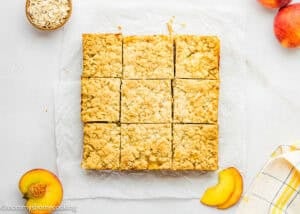 Nine Eggless Peach Crumb Bars are arranged in a square on parchment paper, surrounded by fresh peach slices, a bowl of oats, and a yellow plaid napkin.