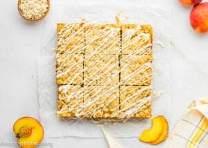 A tray of Eggless Peach Crumb Bars drizzled with white icing, cut into squares, is displayed on parchment paper with peach slices and oatmeal nearby.