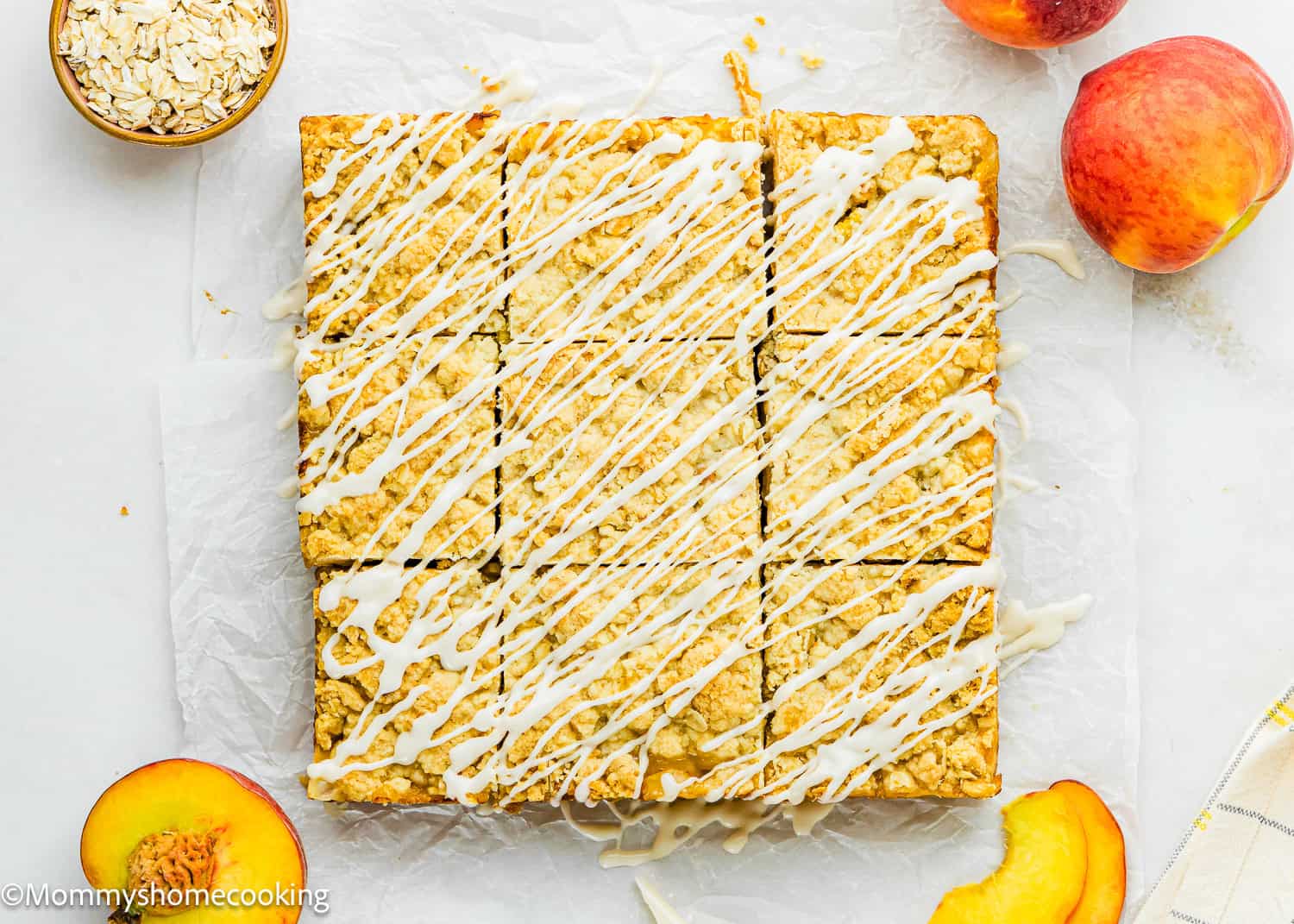 Nine square Eggless Peach Crumb Bars with white icing drizzle, surrounded by fresh peaches, peach slices, and a bowl of oats on a white surface.